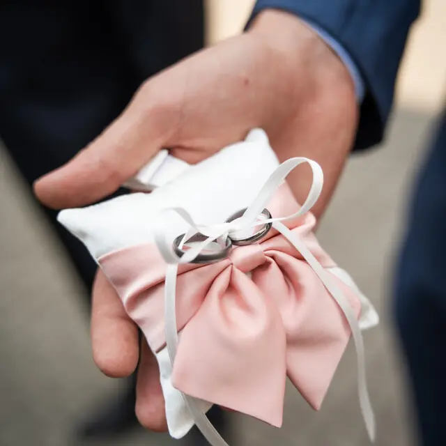 En kudde med en rosa fjärilsrosett, med vigselringar uppbundna med ett vitt organzaband, i brudgummens hand.