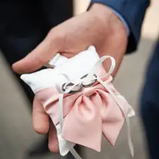 En kudde med en rosa fjärilsrosett, med vigselringar uppbundna med ett vitt organzaband, i brudgummens hand.